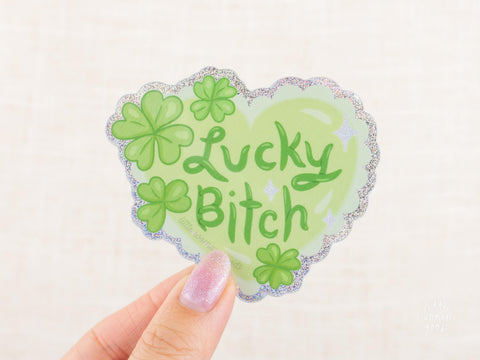 A glittery, green heart sticker with the text "lucky bitch". Four leaf clovers adorn the heart.